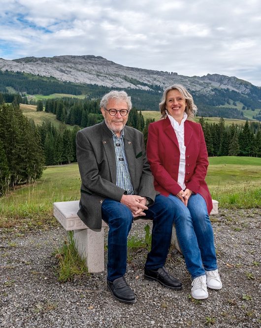 Foto von den Gastgebern vom Hotel Bergwelten Salwideli. Sie sitzen auf einer Bank und im Hintergrund ist die wundervolle Natur vom Sörenberg zusehen.